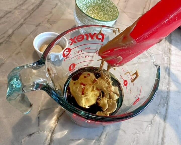 Red spatula over a clear measuring cup with brown liquid and sunflower seed butter.