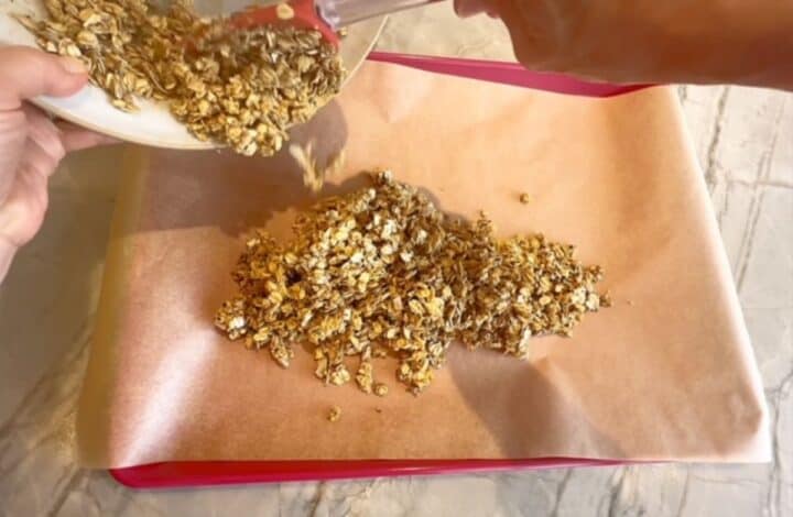 Pouring raw granola onto a red-pink baking sheet lined with parchment paper.