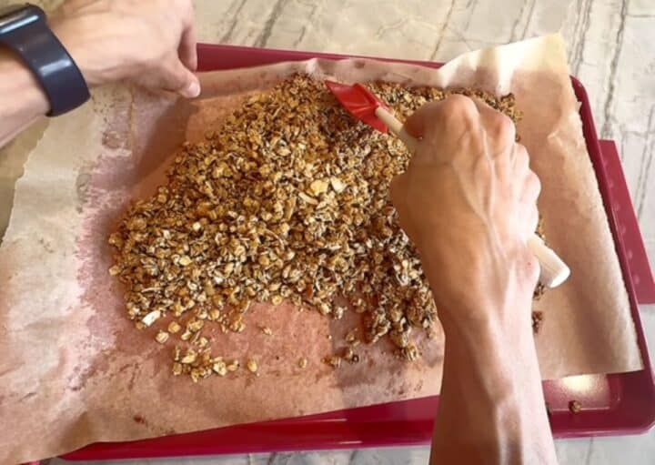 TRight Hand using red spatula to stir baked granola and left hand picking up parchment on baking sheet