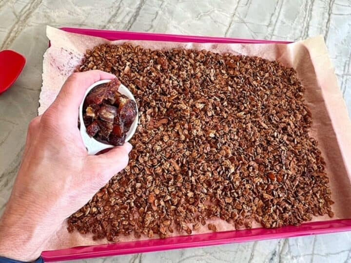 Left hand holding a white container full of dates over golden brown granola on a sheet pan.