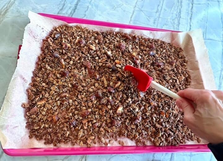 Right hand using red spatula to stir chopped dates into golden brown granola on sheet pan.