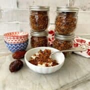 White fluted bowl with plain yogurt topped with granola and jars of granola behind it plus whole dates beside it.