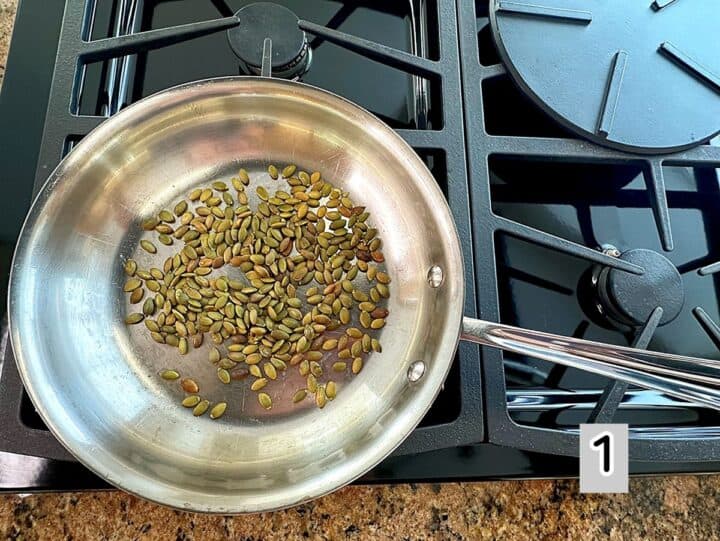 Aluminum skillet on black grate of gas cooktop toasting greenish pumpkin seeds.
