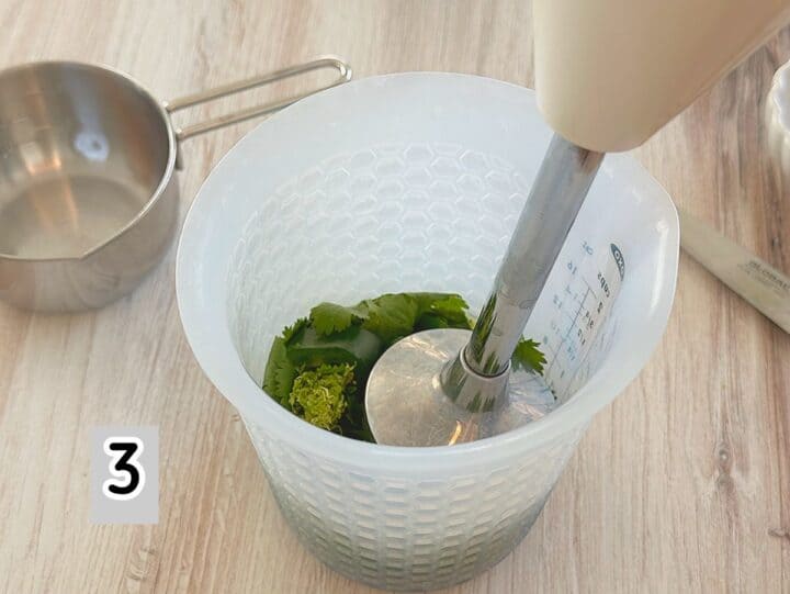Stainless head of stick blender inserted into green herbs and seeds in an opaque blender container.