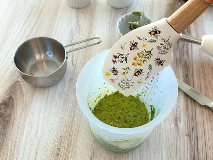 Spatula decorated with bees going into an opaque silicone blender container filled with green sauce on a wooden background.