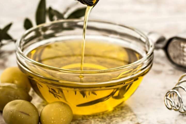Clear bowl full of yellow-green olive oil surrounded by green olives and point leaves