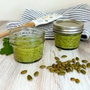Two small (4-oz) jars of thick green sauce with pumpkin seeds in front and a spatula and blue-striped kitchen towel in the back.