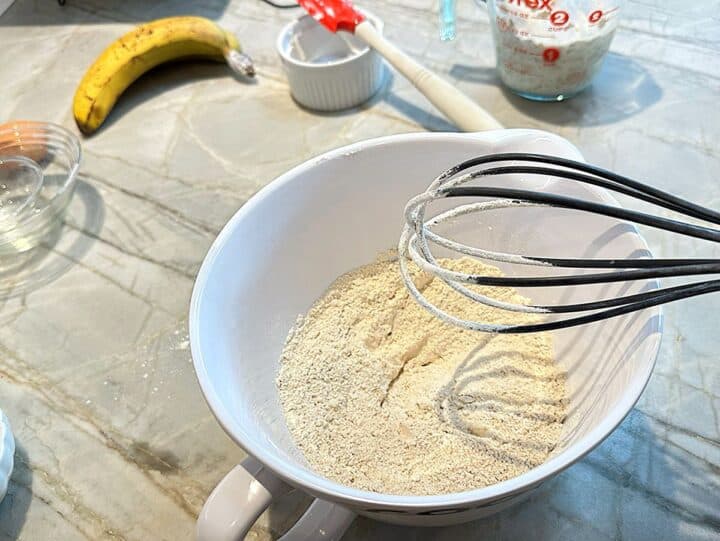 Black silicone whisk hovering over white bowl with flour and dry ingredients on marble counter with banana in the background.