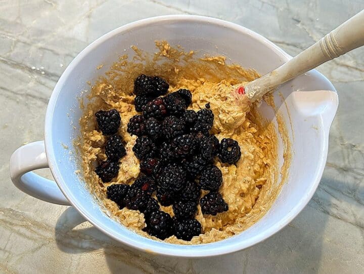 A mound of blackberries sit atop muffin batter in a white bowl on a marble countertop. 