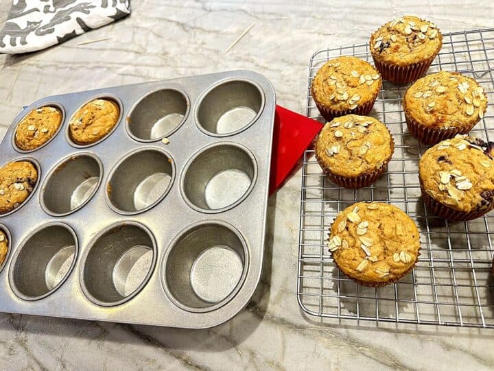 Gray muffin tin with a oat-sprinkled muffin in it next to a wire rack with muffins cooling on top. 