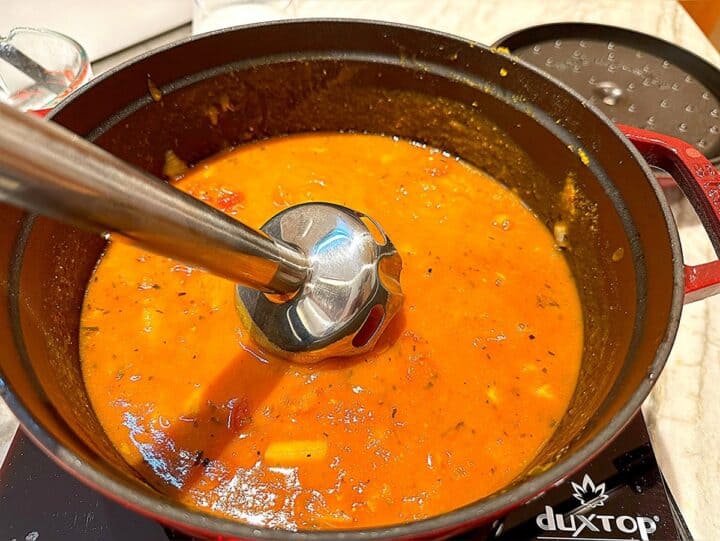 Silver immersion blender in an orange-colored soup in a black pot.
