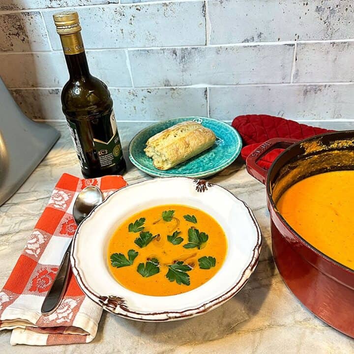 A white bowl or orange-colored pumpkin soup with parsley leaves on top plus a small blue-green plate with a hunk of bread and a vial of olive oil.