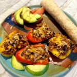 Red and yellow bell pepper quarters with stuffing and melted cheese plus avocado-grapefruit salad and tortilla on a plate.