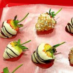 Four white-chocolate-covered strawberries with dark chocolate and pink-gold sugar decorations.
