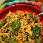 Creamy, curried chickpea rotini with chicken and spinach in a flat, red pasta bowl against a towel with a blue-green Southwestern pattern