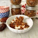 White bowl with white yogurt topped with brown granola with jars of granola behind it.
