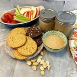 Oval dish with slices of apple, red pepper, and celery and plate of dark and light brown crackers sits next to jars of tan nut butter and a round dish of tan nut butter on a marble countertop.